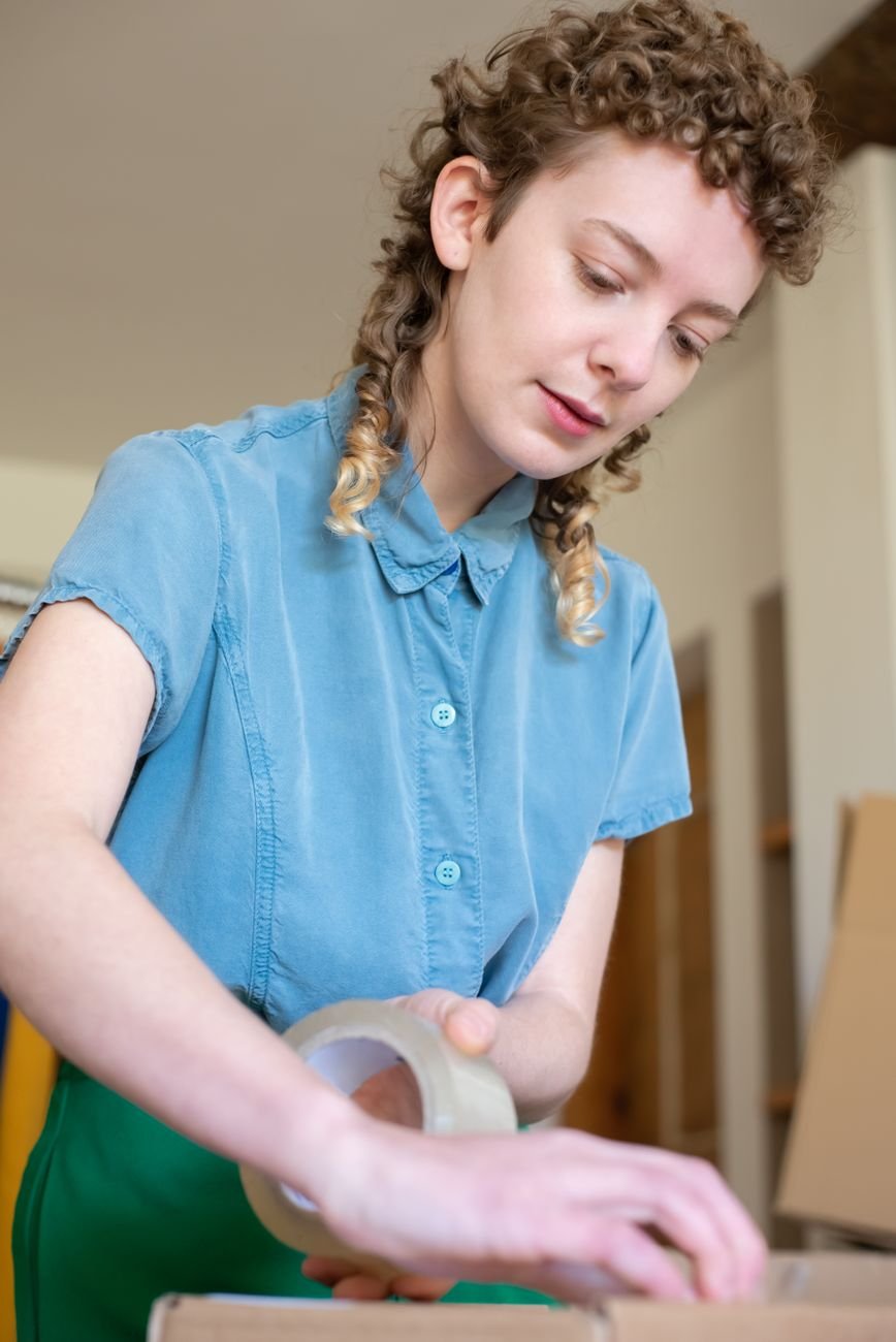 Young woman with curly hair using tape for packaging indoors.